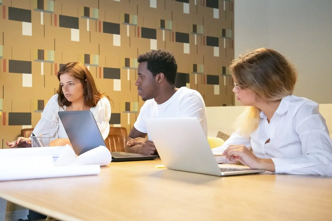 Eine vielfältige Gruppe von Designern arbeitet gemeinsam an einem Projekt, sitzt an einem Tisch mit Laptops und Blaupausen und spricht miteinander.